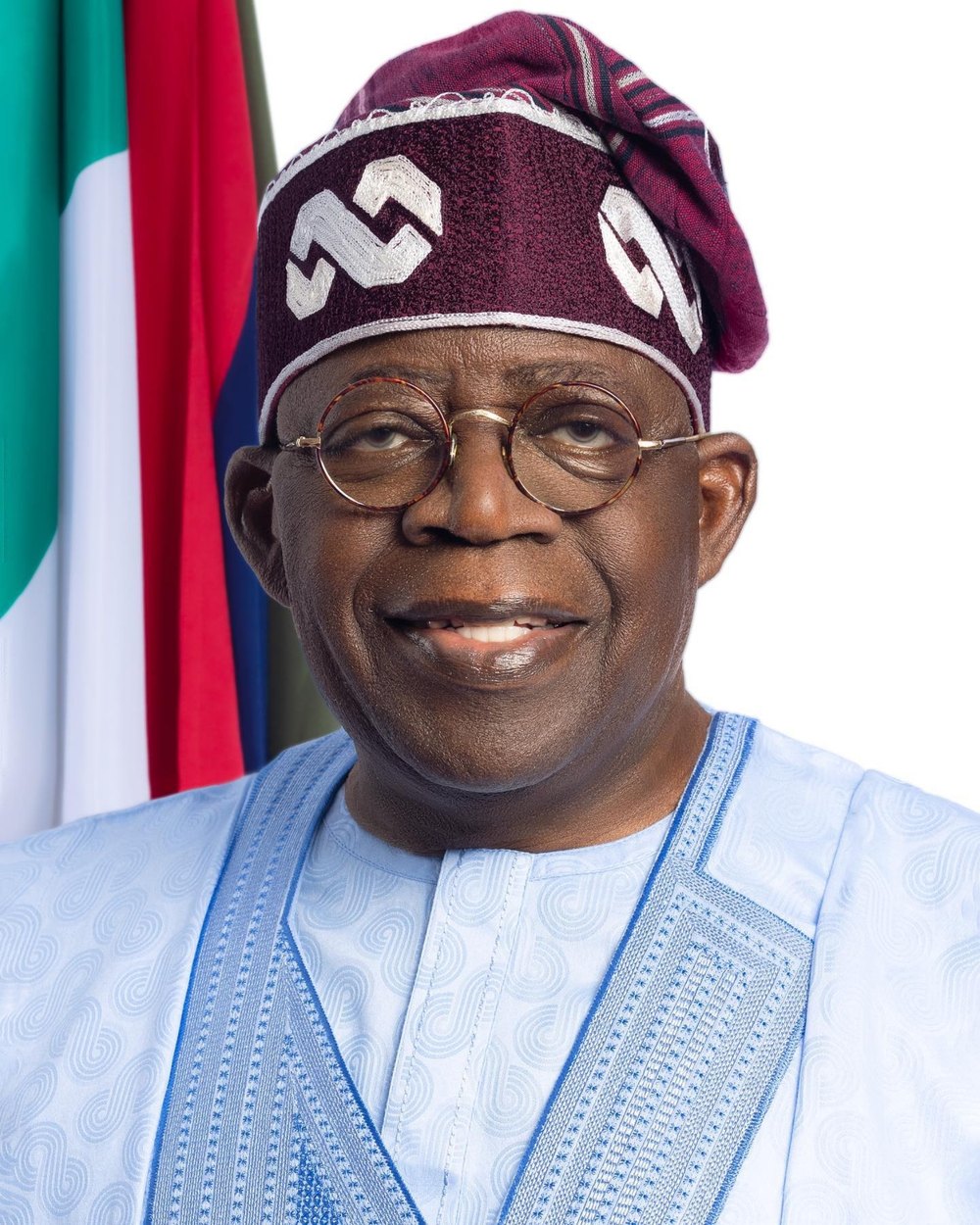 Official portrait of His Excellency Bola Ahmed Tinubu, GCFR, President of the Federal Republic of Nigeria, smiling, in a maroon embroidered cap, round glasses and light blue attire, with the Nigerian flag behind him